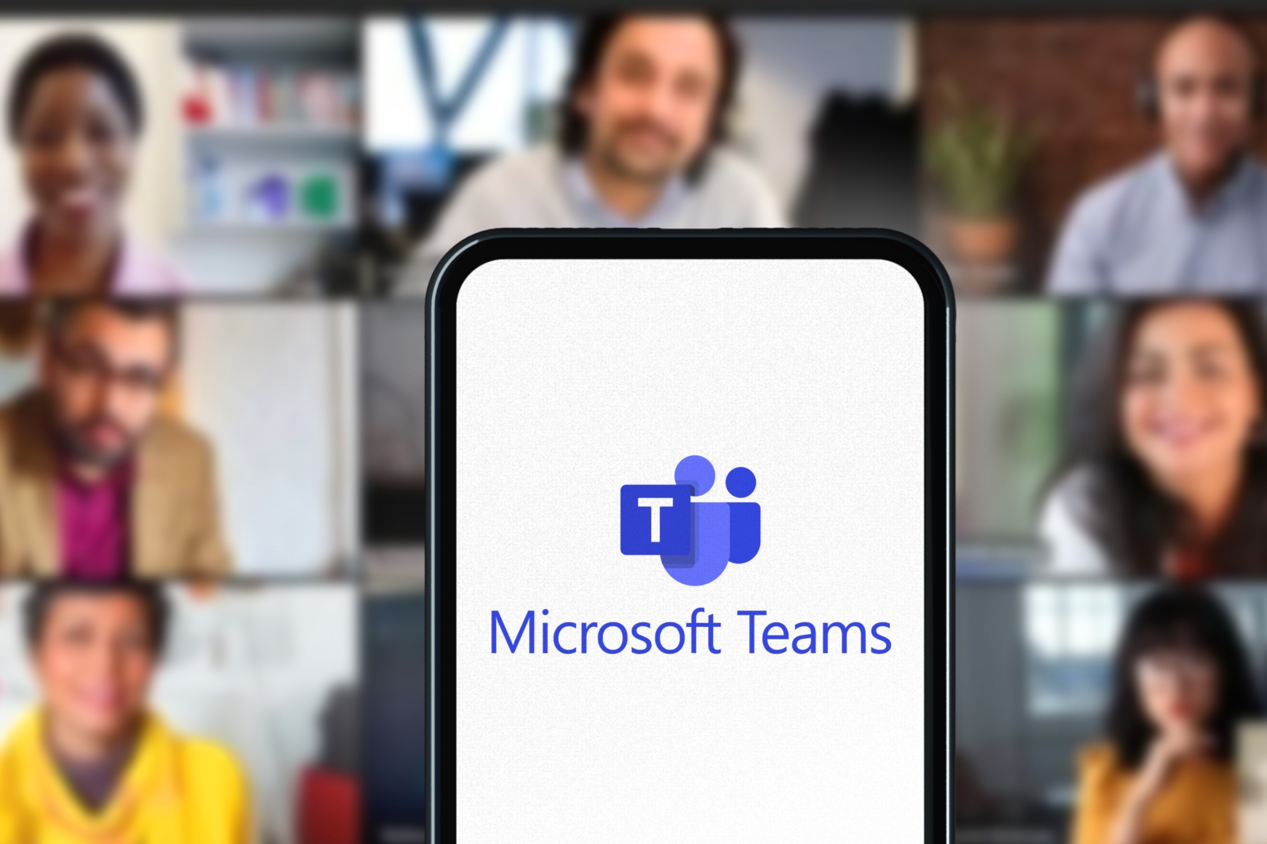 A phone showing the Microsoft Teams Logo is set in front of a background of individuals all on a video call.